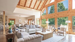 Ein großzügiges Wohnzimmer mit einer großen Couch, einem Couchtisch und Sesseln, Holzbalken an der Decke und großen Fenstern mit Blick ins Freie.