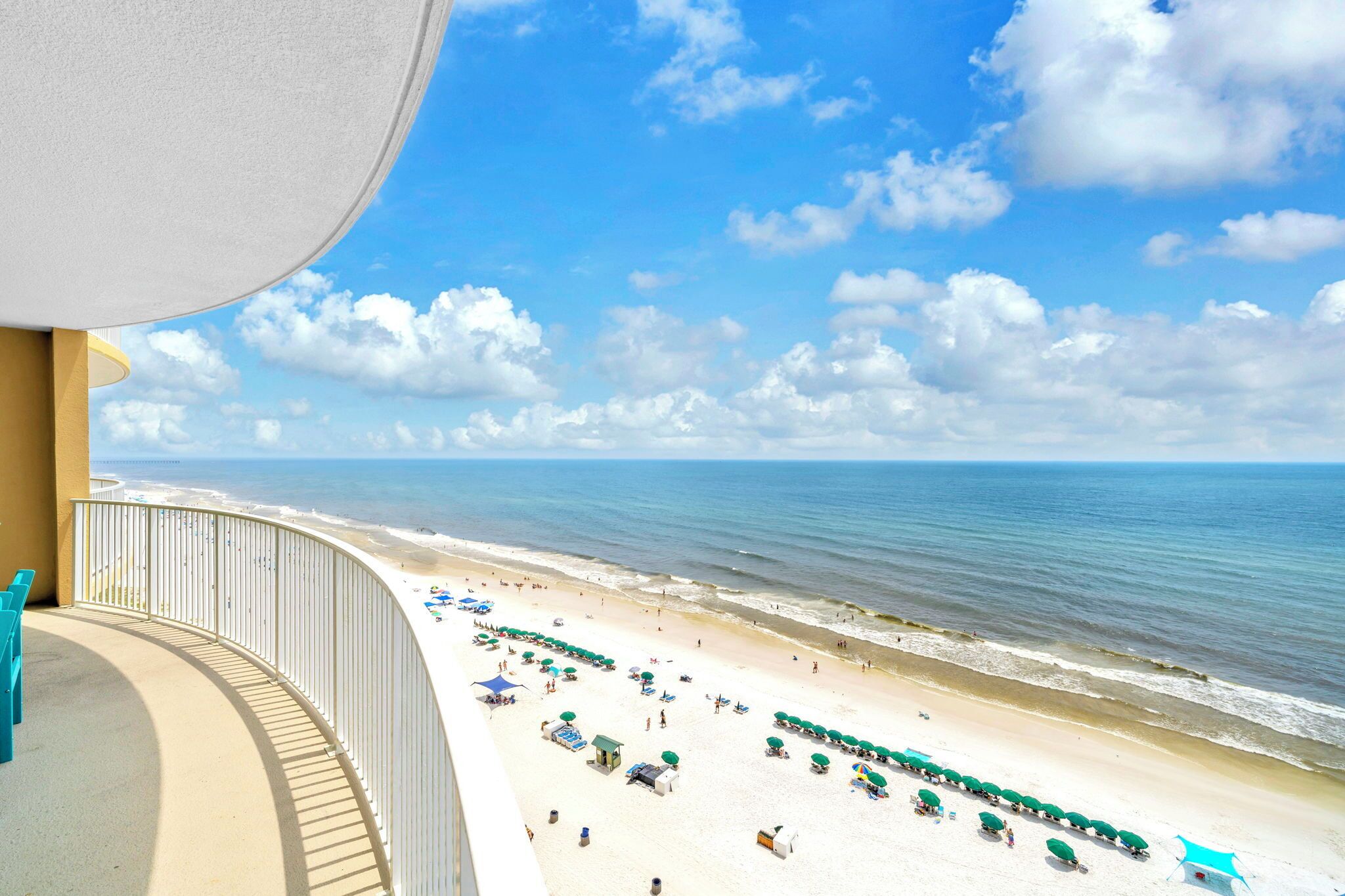 Blick vom Balkon auf einen Strand mit einer Geländer, feinem Sand, Sonnenschirmen und Liegestühlen, sowie dem Meer im Hintergrund.