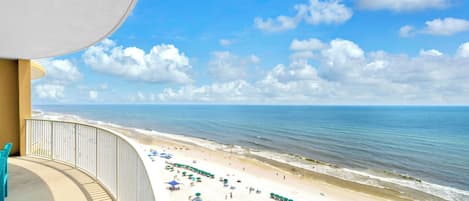 A beach view from a balcony with a railing, a sandy beach with umbrellas and lounge chairs, and the ocean in the background.