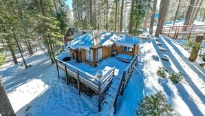 A wooden cabin with a deck, surrounded by snow-covered ground and trees.