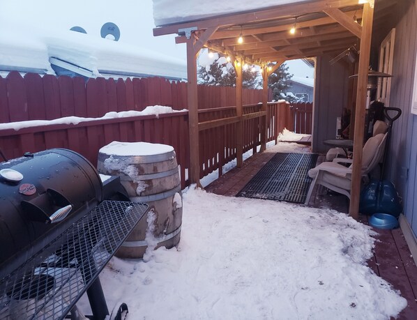 A covered outdoor area with a barbecue grill, a metal barrel, and a wooden deck.