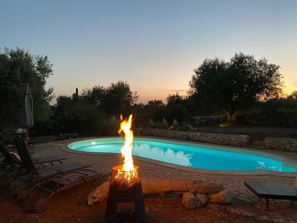 A swimming pool with a fire feature in the foreground, surrounded by lounge chairs and a stone wall. - A romantic trullo with a private pool surrounded by nature (Ceglie Messapica)