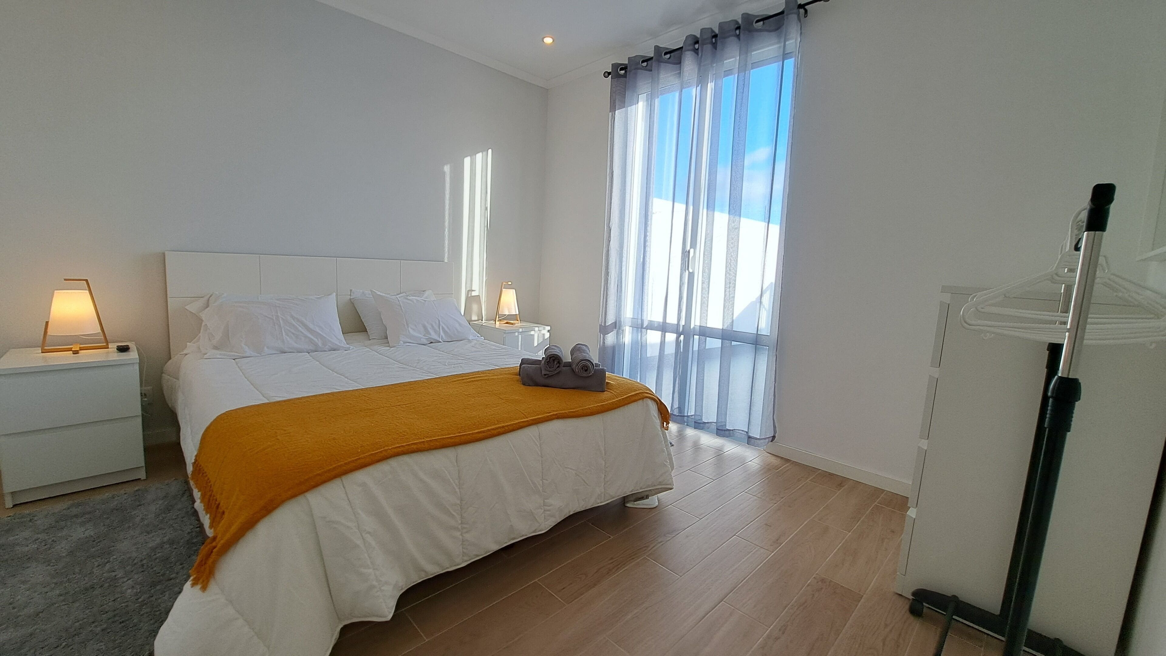 Ein modernes Schlafzimmer mit einem großen Bett, weißer Bettwäsche, Holzboden, einem Nachttisch mit Lampe und einem Fenster mit durchsichtigen Vorhängen.