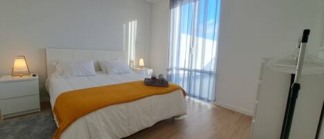 Un dormitorio moderno con una cama grande, ropa de cama blanca, piso de madera, una mesita de noche con una lámpara y una ventana con cortinas traslúcidas.