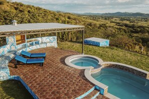 Relax in a serene outdoor oasis with a hexagonal tile patio, shaded seating area, and a hot tub surrounded by a pool.