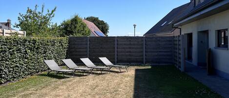 A residential backyard with a wooden fence, a lawn with sun loungers, and a house with a sloped roof.