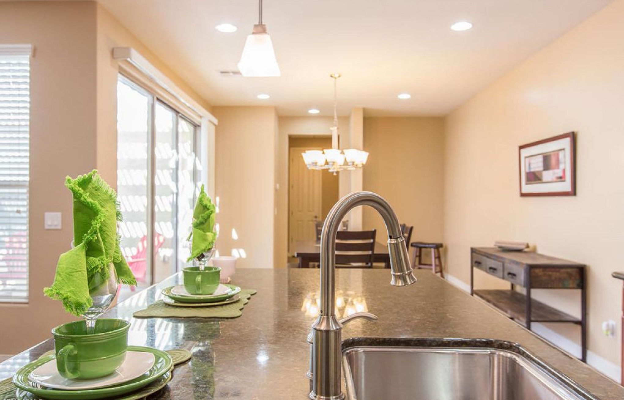 A modern kitchen with a stainless steel sink, a dining area with a chandelier, and a view of the dining room through a large window.
