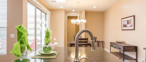 A modern kitchen with a stainless steel sink, a dining area with a chandelier, and a view of the dining room through a large window.