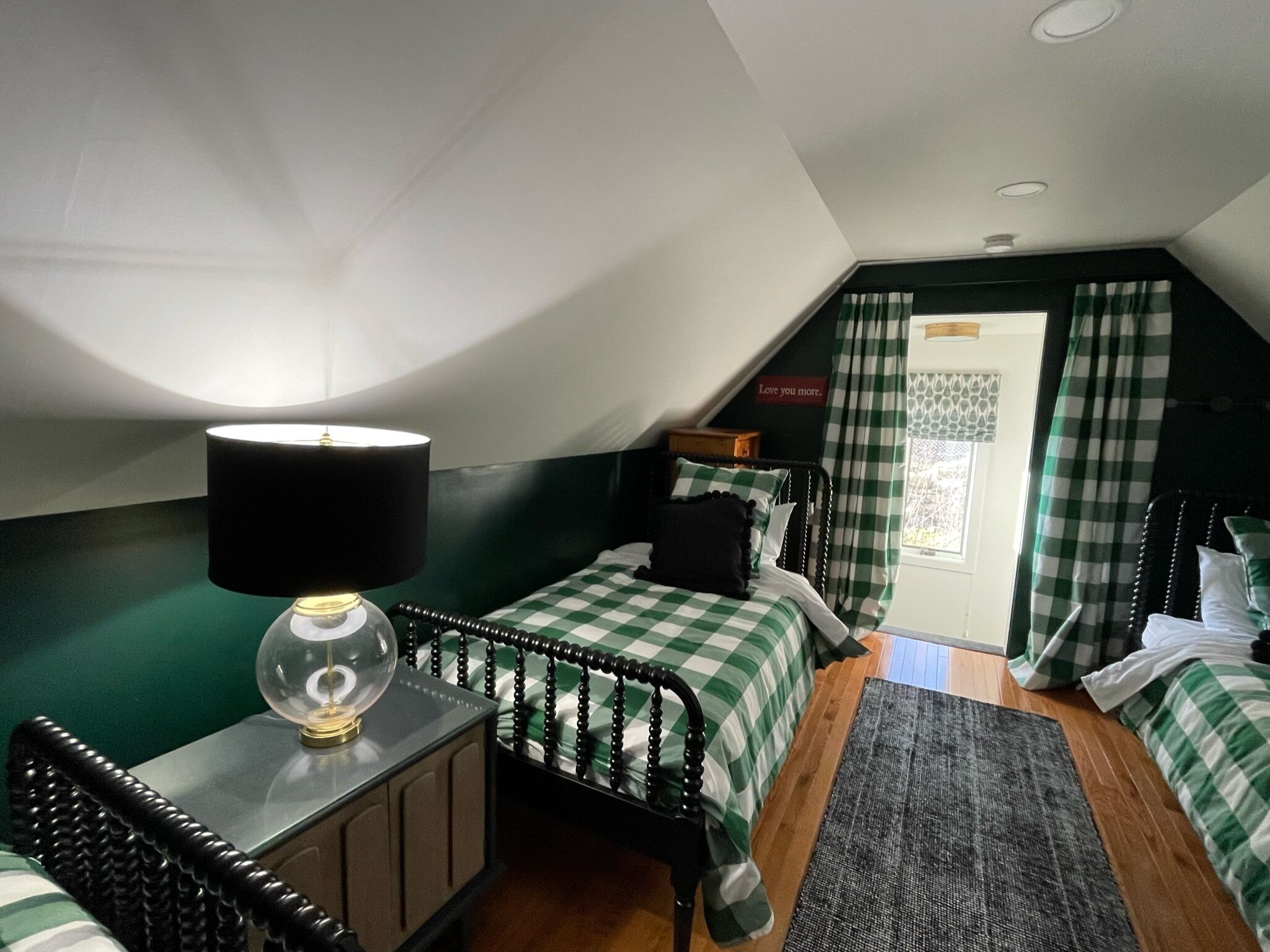 A bedroom with a checkered bedspread, a bedside lamp, a small wooden nightstand, and a view of the outdoors through a window with checkered curtains.