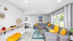 A modern living room with a grey sofa, yellow cushions, a wooden bench, and a dining area in the background.