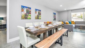 A modern dining area with a wooden table set for six, white chairs, and two paintings on the wall.