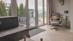 A modern living room with a large sliding glass door leading to a balcony, a comfortable armchair, a small table with a lamp, and a mounted deer skull on the wall. - Penthouse Lake Mountains View Incl Summer Card (Zell am See)