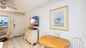 Ein Zimmer mit Holztisch, weißen Stühlen, Deckenventilator, Fernseher auf einer Kommode und einem gerahmten Gemälde an der Wand.