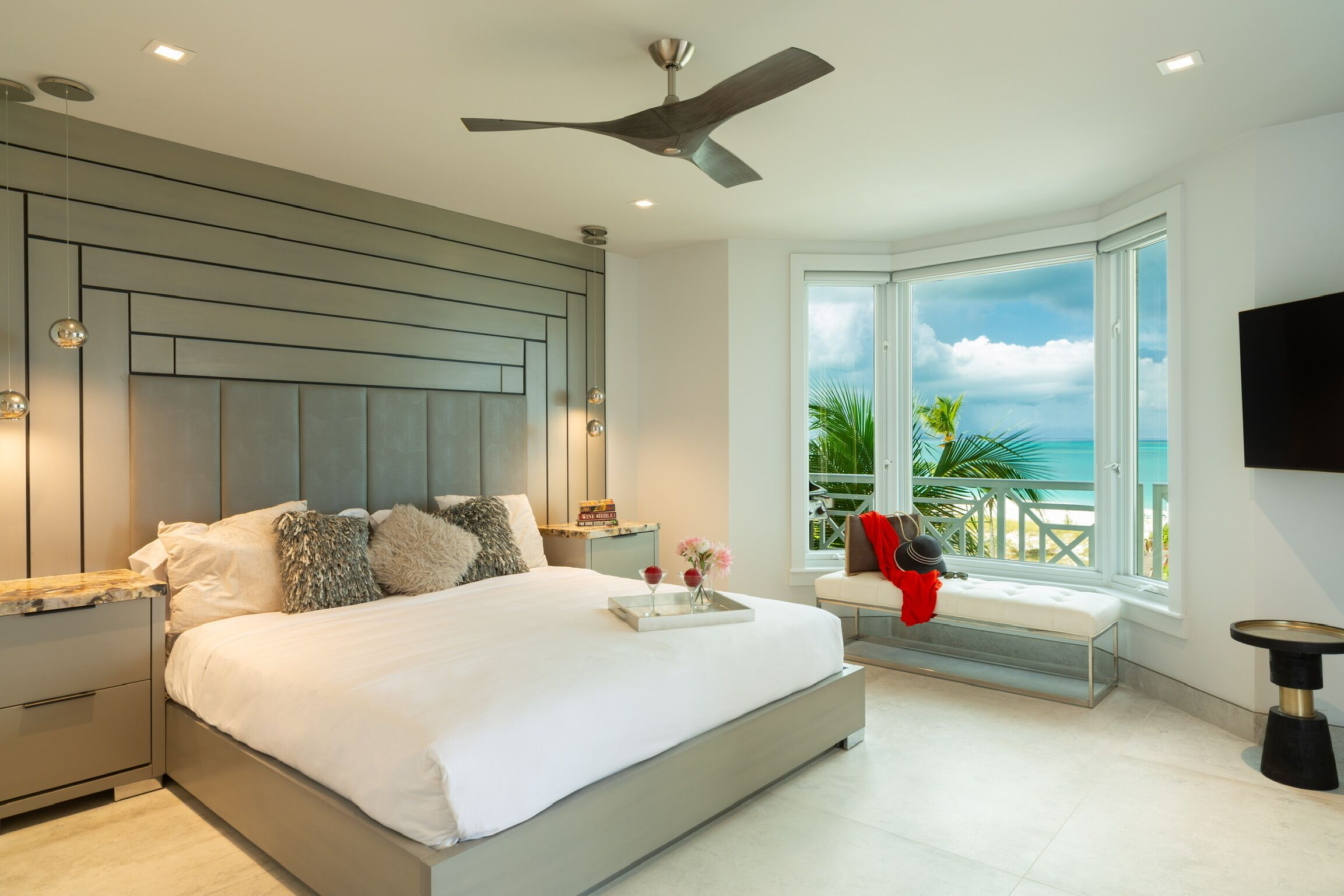 A modern bedroom with a large bed, a ceiling fan, a nightstand, a bedside table, a window with a view of the ocean, and a TV mounted on the wall.