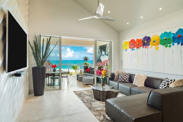 A modern living room with a large sofa, a coffee table, and a TV mounted on the wall. There is a large window with a view of the ocean. - The Blu2 On the Beach, Luxury Oceanfront Penthouse Condo at the Grandview (Grace bay)