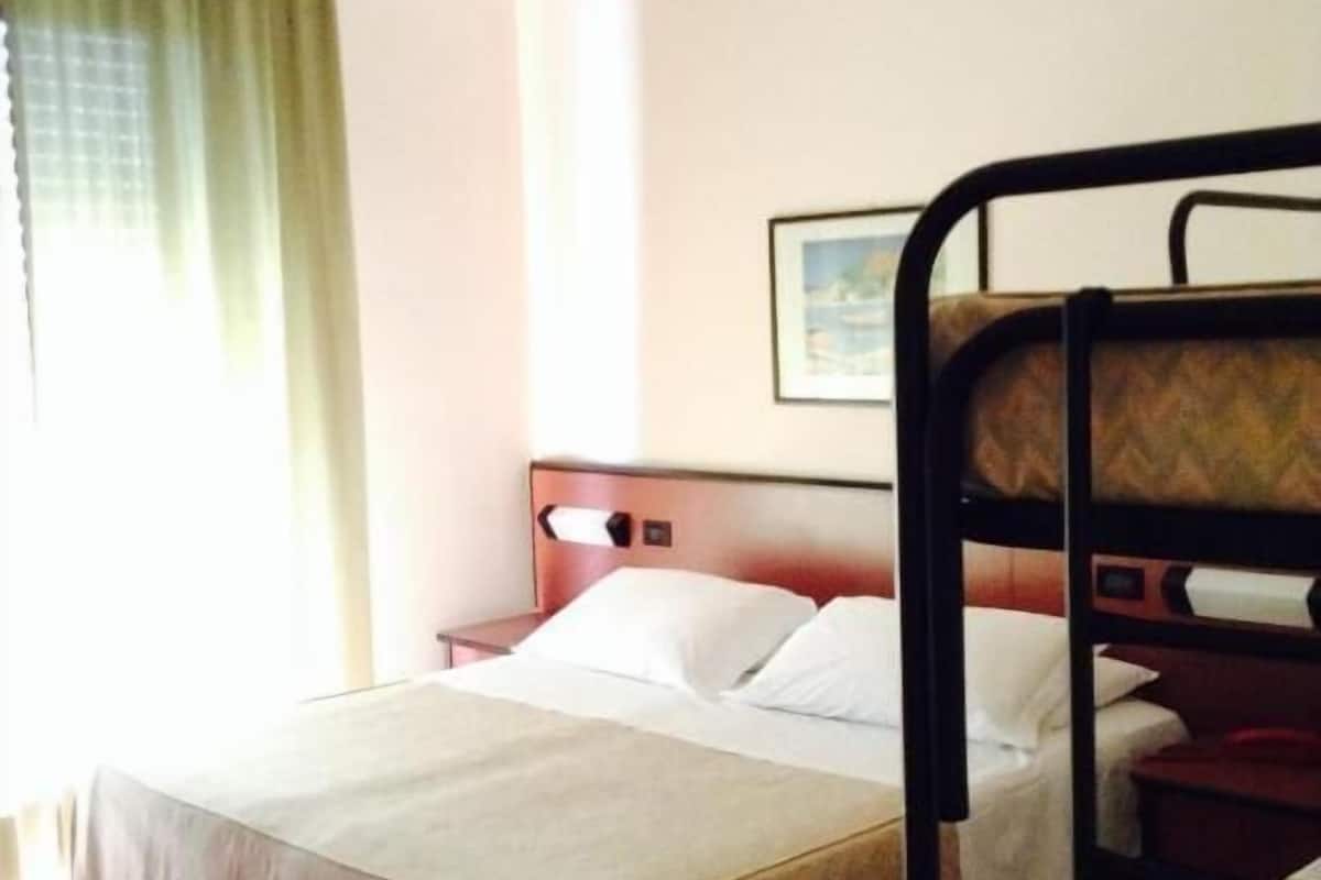 Un lit simple avec une tête de lit en bois et une table de chevet, une fenêtre avec des rideaux et un tableau encadré au mur.