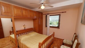 A bedroom with a wooden bed, a ceiling fan, wooden cabinets, a window with curtains, and a chair. - Seaside Apartments Markota - Two Bedroom Apartment with Terrace and Sea View (Prižba)