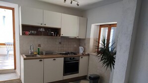 Cocina pequeña con armarios blancos, estufa a gas y lavamanos. Hay un cubo de basura y una planta en maceta cerca de la ventana.