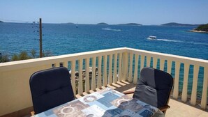 Un balcón con dos sillas y una mesa, con vista a un mar azul cristalino con barcos y islas lejanas.
