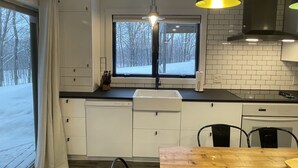A modern kitchen with white cabinets, a black countertop, a window with a view of a snowy landscape, and a wooden dining table. - Ski in/out Condo Modern Mountain Top at Snowriver Resort (Wakefield)