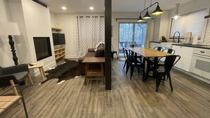 A modern living room with a fireplace, a dining area with a wooden table and chairs, and a kitchen with white cabinets and a window. - Ski in/out Condo Modern Mountain Top at Snowriver Resort (Wakefield)