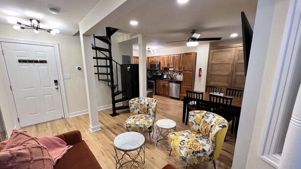 A modern living room with a brown sofa, two floral armchairs, and a small round table. A staircase leads to a kitchen area with wooden cabinets and a microwave.