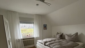 A bedroom with a bed, a radiator, a window with curtains, and a ceiling fan. - Quiet vacation apartment in the beautiful Sauerland (Meschede)