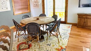 Un coin repas avec une table ronde en bois entourée de six chaises, un tapis à motifs et une fenêtre avec des stores.