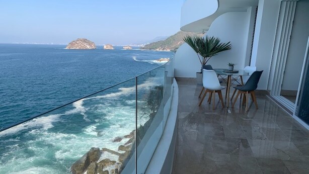 Un balcon doté d’une balustrade en verre donnant sur la mer, un coin repas avec des chaises et une table, et une plante en pot.