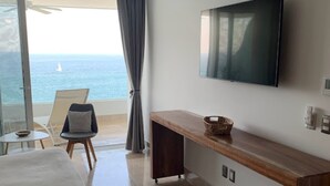 Une chambre d’hôtel avec un lit, un téléviseur fixé au mur, une console en bois, une chaise et une vue sur l’océan par une grande fenêtre.