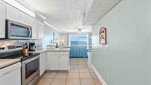 A kitchen with white cabinets, a microwave, and a stove. There is a dining area with a blue-striped tablecloth and a view of the ocean.