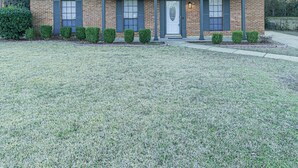 A single-story brick house with a dark roof, a front door, and a small front yard with trimmed bushes.