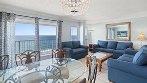 A living room with a glass table, blue sofas, a chandelier, and a view of the ocean. - The Summit PCB 1504 ** Large Updated Condo with Huge Beachfront Balcony (Panama City Beach)