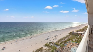 Uitzicht op een strand vanaf een balkon, met een heldere lucht, een zandstrand en enkele gebouwen in de verte.