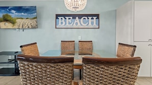 Een eethoek met een glazen tafel en geweven stoelen, een kroonluchter, een bord met 'BEACH' erop en een schilderij van een strandgezicht.