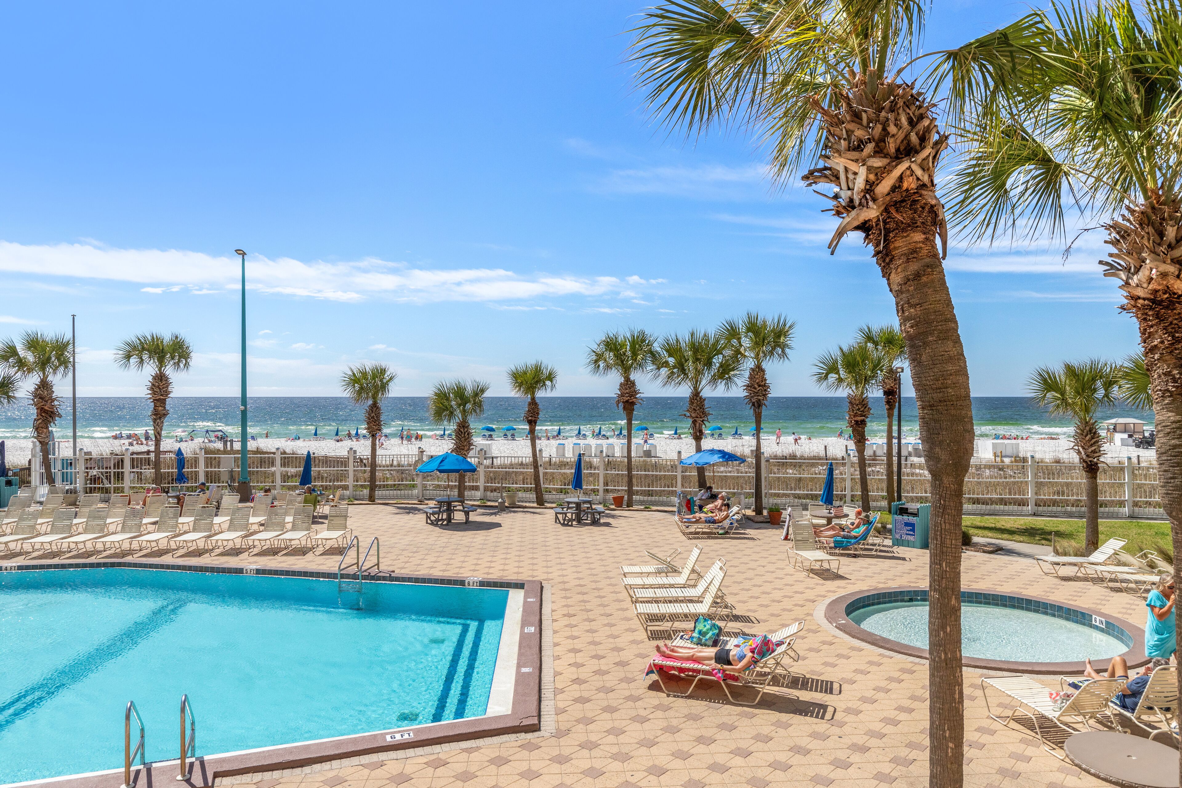 A pool area with lounge chairs and umbrellas, a beach in the background, and a clear sky.