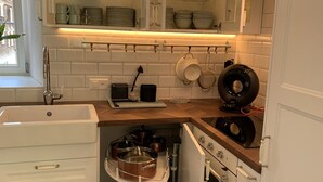 A compact kitchen with white cabinets, a built-in oven, and a sink. There is a window providing natural light, and a shelf above the sink holding dishes and glasses. - Allegra Appenzell (Trogen)