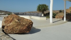 Ein moderner Sitzbereich im Freien mit einem großen Felsen als Mittelpunkt, einer weißen Säule und Blick auf die Küste.