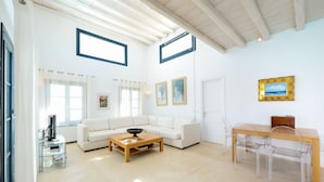 Ein modernes Wohnzimmer mit einem weißen Sofa, einem Holztisch, einem Glastisch mit Stühlen und zwei gerahmten Bildern an der Wand. Ein Fenster mit Vorhängen ist ebenfalls vorhanden.