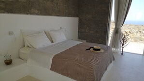 Ein Schlafzimmer mit Steinwand, einem Bett mit weißer Bettwäsche, einer braunen Decke und Blick ins Freie durch ein Fenster.