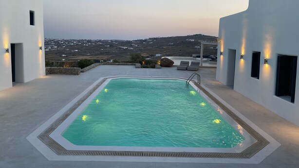 Ein rechteckiger Pool mit Steinrand, umgeben von weißen Wänden mit kleinen Fenstern und Wandleuchten.