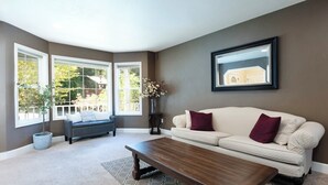 A living room with a dark wooden coffee table, a cream-colored sofa with maroon pillows, a dark sofa, and a large window with a view of greenery. - Sopra Bella- Elegant 4BR, Family Friendly, OSU (Corvallis)