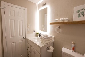 Une salle de bain avec une porte blanche, un lavabo surmonté d’une pile de serviettes, un miroir et des toilettes avec un porte-rouleau de papier toilette.