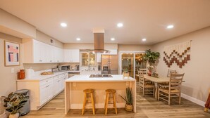 Una cocina moderna con armarios blancos, una isla central con taburetes, un comedor con sillas de madera y una planta grande en maceta.
