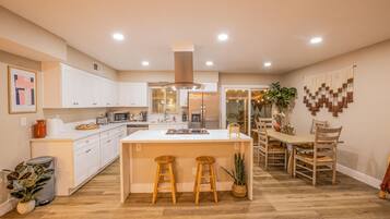 Una cocina moderna con armarios blancos, una isla central con taburetes, un comedor con sillas de madera y una planta grande en maceta.