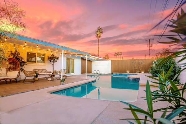 A pool area with lounge chairs and string lights, surrounded by plants and a sunset sky.
