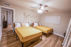 Une chambre avec deux lits, un ventilateur de plafond, une fenêtre avec des stores et une porte donnant sur une autre pièce.
