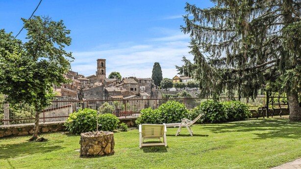 Una zona exterior amb gespa, amb una banca de fusta, una jardineria de pedra i una tanca metàl·lica. Al fons, es pot veure una ciutat antiga amb edificis i una torre.