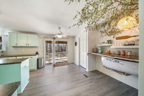 Una cocina con gabinetes verde claro, encimera blanca y suelo de madera. Hay un ventilador de techo, una puerta que da al exterior y una estantería con frascos y un bate de béisbol.