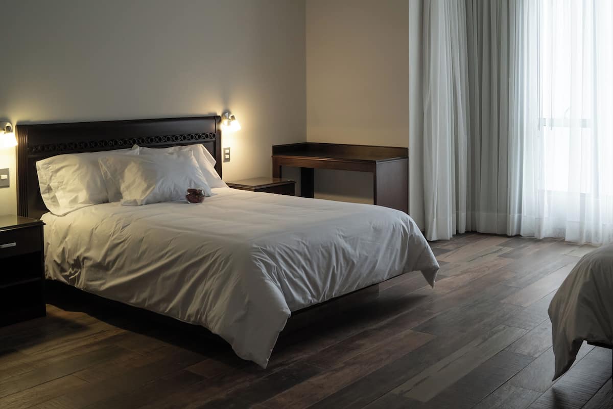 Ein Hotelzimmer mit einem großen Bett, einem Nachttisch, einem Schreibtisch und einem Fenster mit Vorhängen.